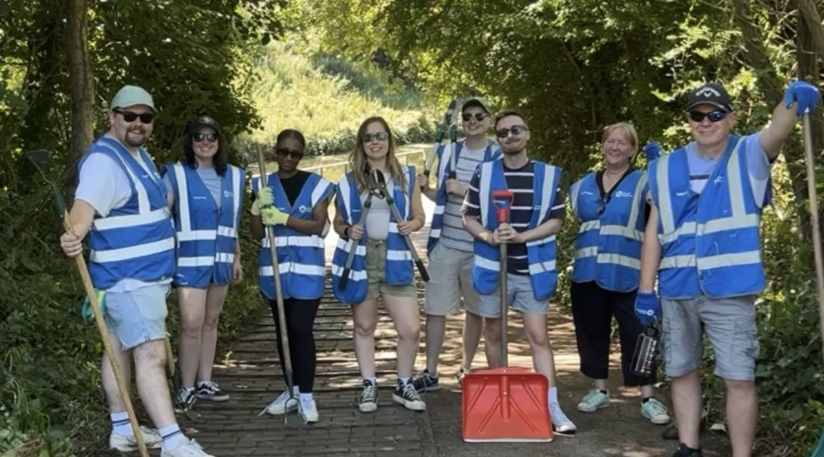 Members of the WPR team in blue high vis jackets, holding rakes and spades, in front of the canal during their canal clean up day